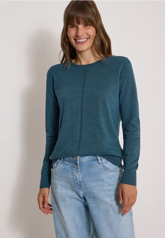 Eine lächelnde Frau in einem petrolfarbenen Langarmshirt und hellen Jeans vor neutralem Hintergrund.