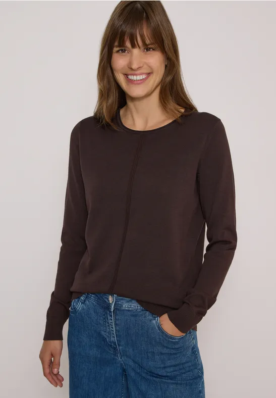 Femme souriante en pull marron et jean devant un fond clair.