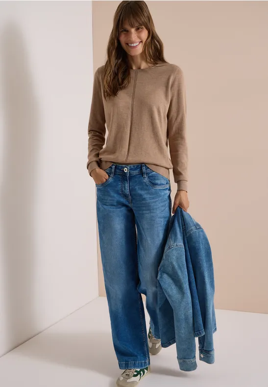 Frau im beigen Cardigan und weiten Jeans, hält eine Jeansjacke in der Hand vor hellem Hintergrund.