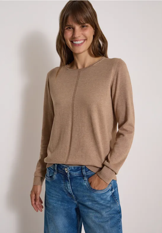 Frau lächelt in hellbraunem Pullover und blauer Jeans, Hände in den Taschen.