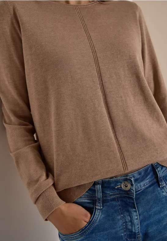 Person trägt braunen Pullover mit Mittelnaht und blaue Jeans, eine Hand in der Hosentasche.