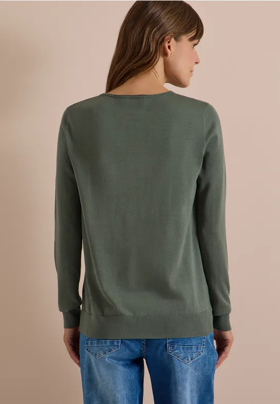 Eine Frau von hinten, trägt einen schlichten grünen Pullover und blaue Jeans vor neutralem Hintergrund.