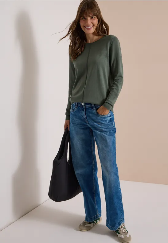 Frau in legerem Outfit: grünes Langarmshirt, lockere Jeans, Sneaker und schwarze Tasche vor weißer Wand.