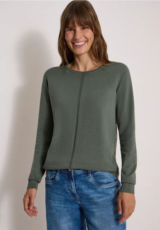 Eine lächelnde Frau in grünem Pullover und blauen Jeans vor hellem Hintergrund.