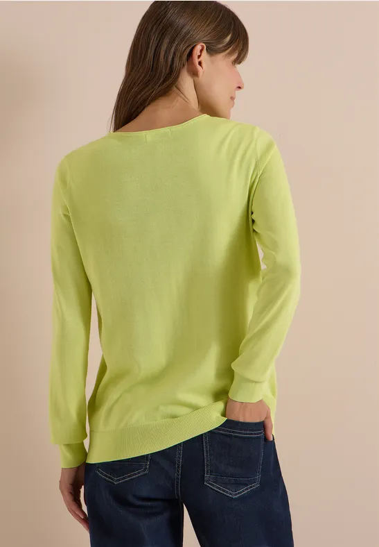 Frau von hinten in neongelbgrünem Langarmshirt und blauen Jeans vor beigem Hintergrund.