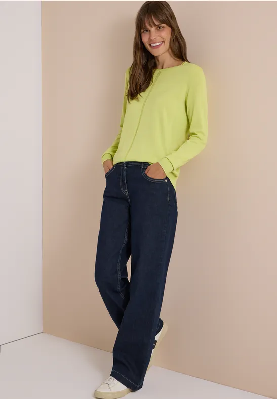 Frau in limettengrünem Pullover, weiten Jeans und weißen Sneakern lehnt lächelnd entspannt an der Wand.