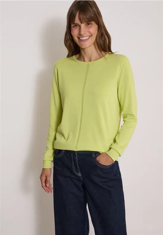Frau im grünen Pullover und blauen Jeans, lächelnd vor hellem Hintergrund.