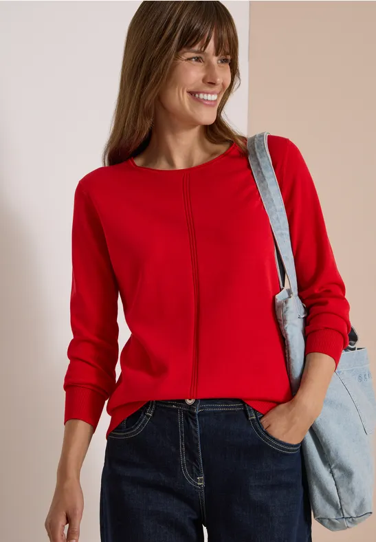 Une femme souriante en pull rouge et jean, portant un sac bleu clair à l'épaule.