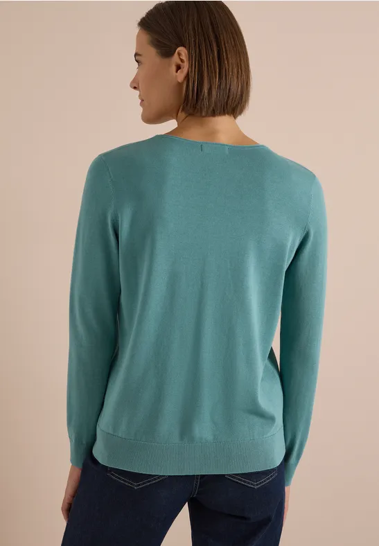 Eine Frau von hinten in einem türkisfarbenen Pullover und Jeans vor neutralem Hintergrund.