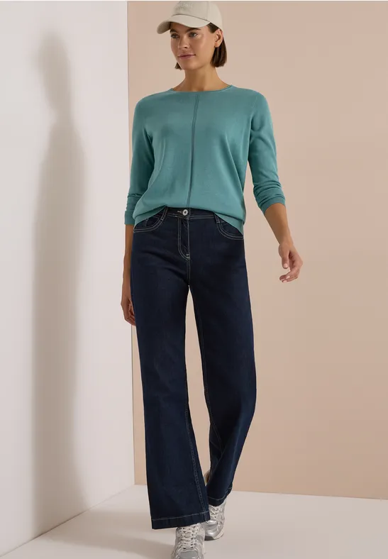 Frau in blaugrünem Langarmshirt, weit geschnittenen Jeans, Sneakern und Cap vor beige Wand.