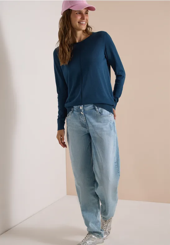 Frau in lockeren hellen Jeans, dunkelblauem Langarmshirt, weißen gemusterten Sneakern und pinker Cap.