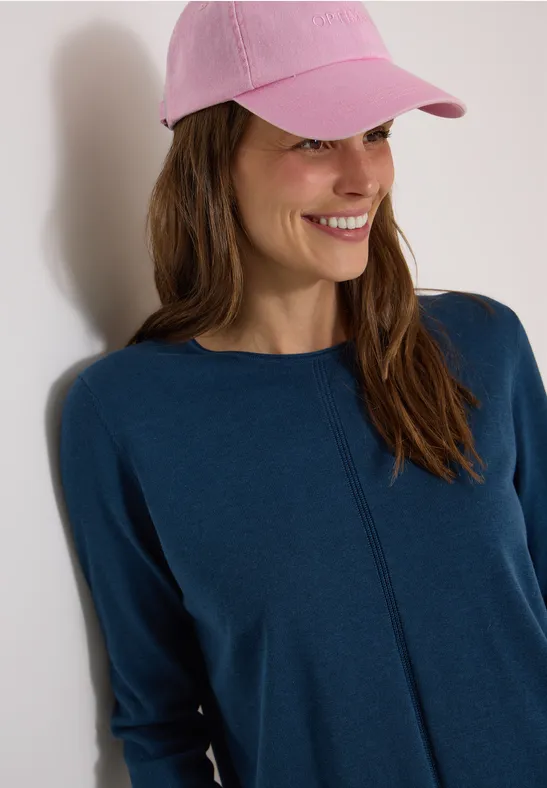 Eine lächelnde Frau mit rosa Cap und blauem Oberteil vor heller Wand.