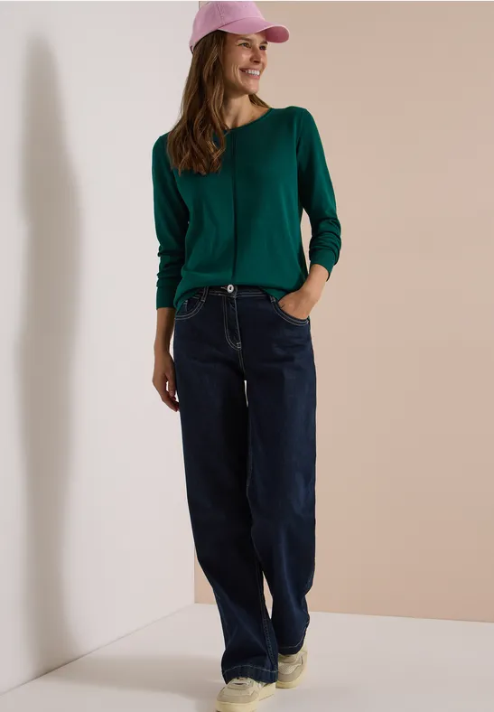 Femme décontractée en pull vert, jean large, baskets beiges et casquette rose, posant contre un mur clair.