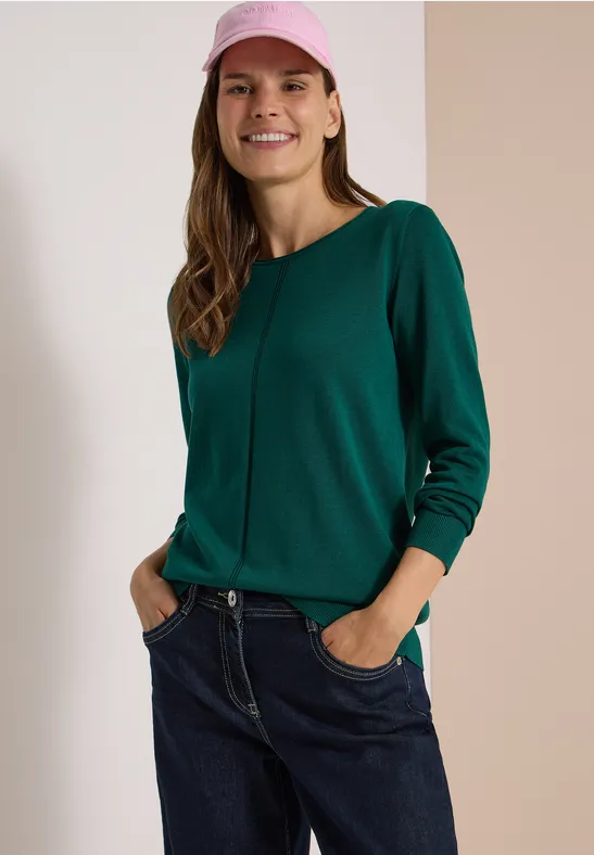Junge Frau in grünem Pullover, Jeans und rosa Mütze, die lächelnd die Hände in den Taschen hält