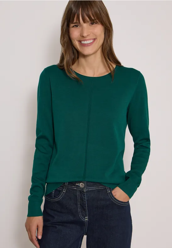 Jeune femme souriante en pull vert et jean devant un fond neutre.