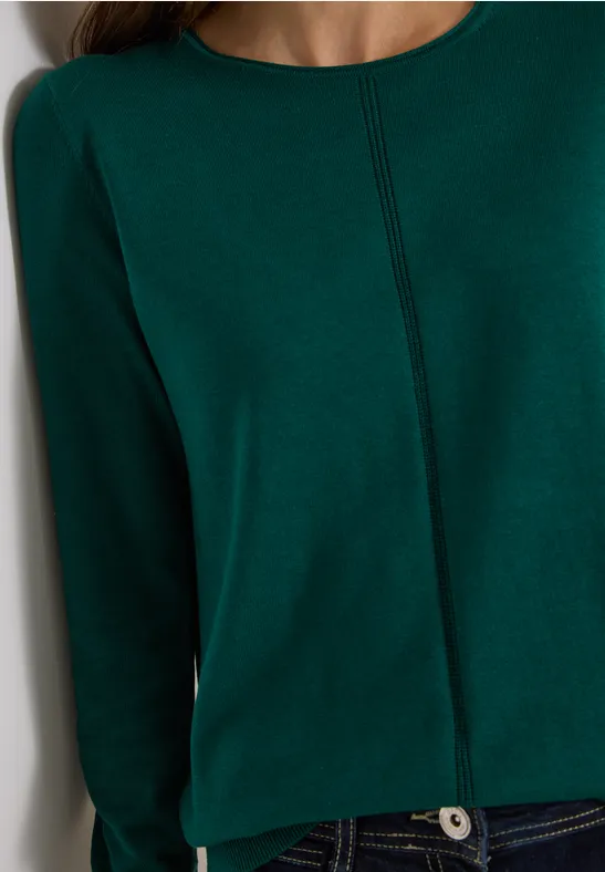 Gros plan sur un pull vert foncé à surpiqûre centrale porté avec un jean bleu.