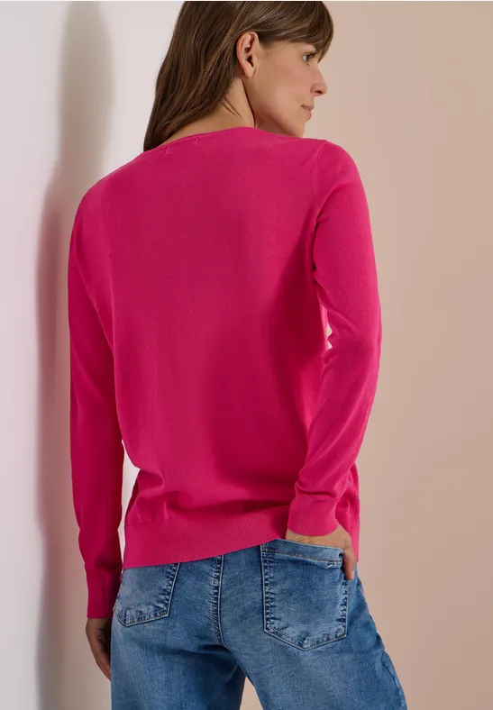 Frau von hinten in pinkem Pullover und blauer Jeans, lehnt an einer hellen Wand.