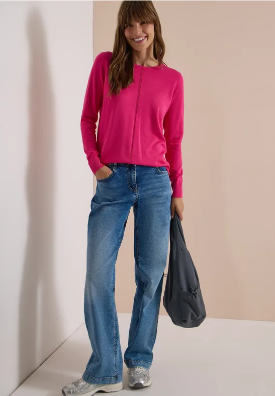 Frau in pinkem Pullover und weitem Jeans mit grauer Tasche und weißen Sneakern lehnt lässig an Wand.