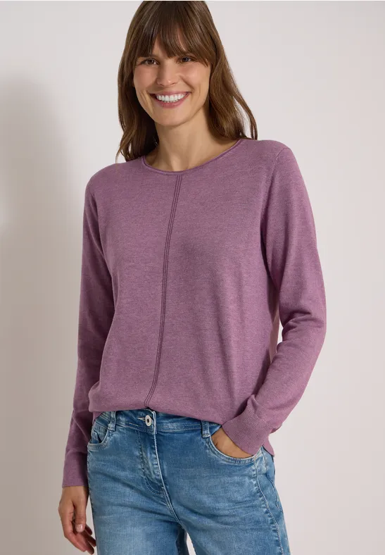 Eine lächelnde Frau in lila Langarmshirt und blauen Jeans vor neutralem Hintergrund.