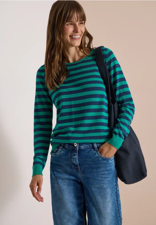 Frau in grün-blau gestreiftem Pullover und Jeans, lächelnd mit Umhängetasche vor neutralem Hintergrund.