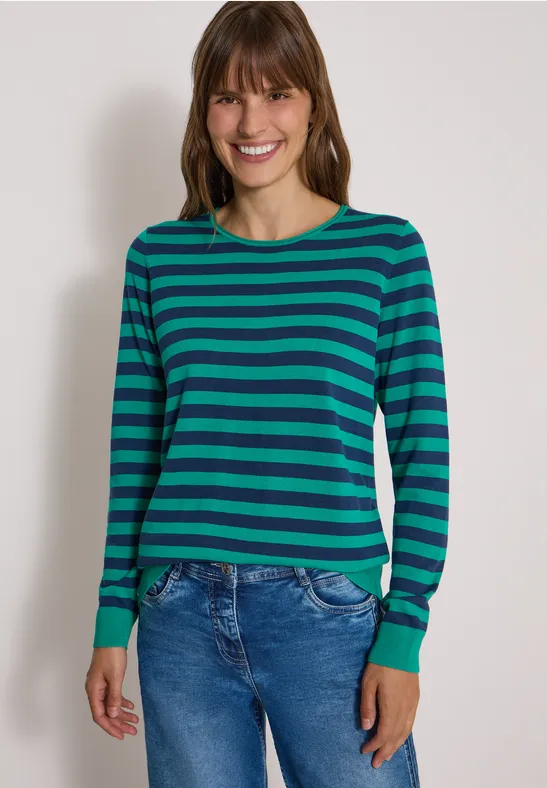 Frau in grün-blau gestreiftem Pullover und Jeans vor neutralem Hintergrund.