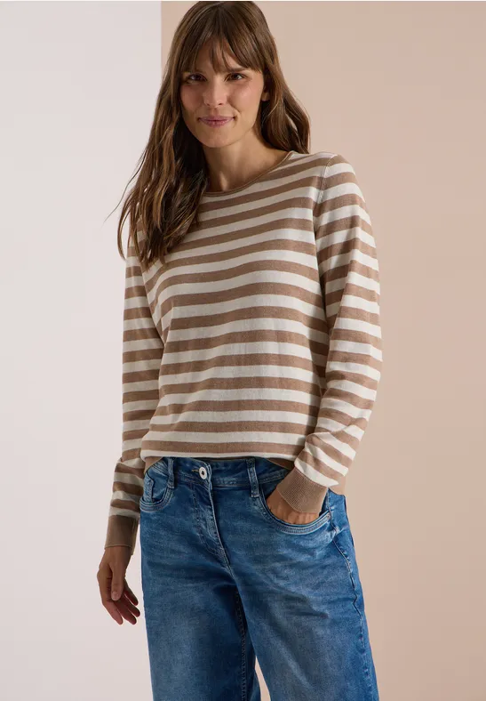Eine Frau in beige-weiß gestreiftem Pullover und blauen Jeans steht lächelnd mit Händen in den Taschen.