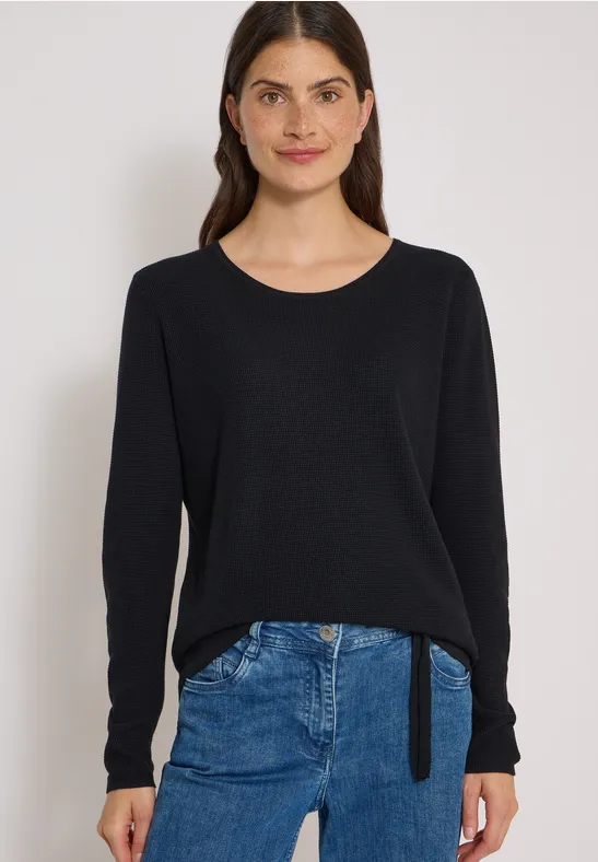 Eine Frau trägt einen schwarzen Pullover und eine blaue Jeans vor hellem Hintergrund.