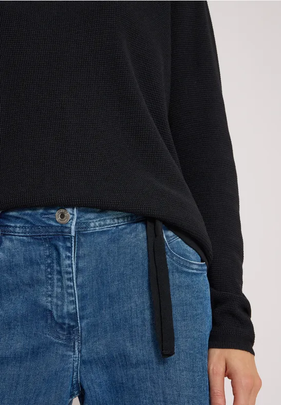 Person trägt schwarzen Pullover und blaue Jeans, nur Oberkörper bis Hüfte sichtbar.