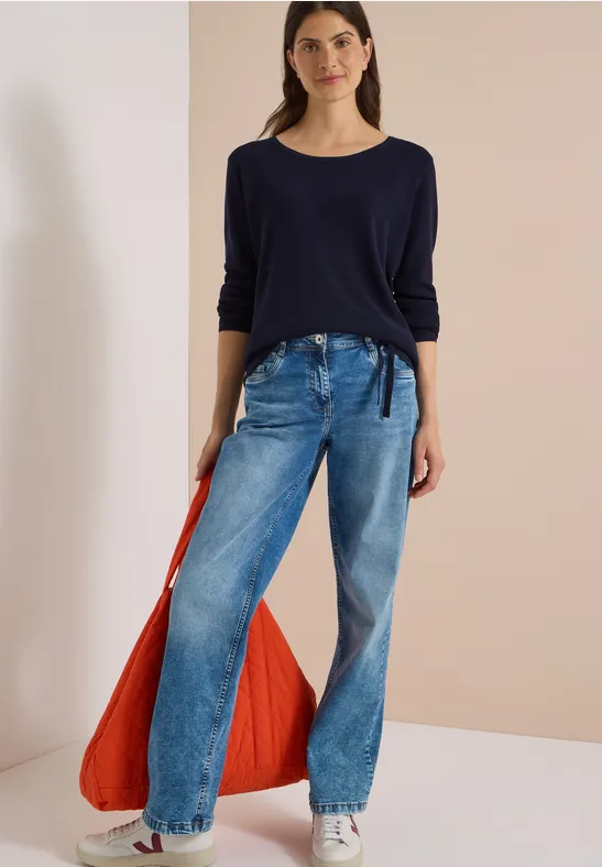 Frau in dunkelblauem Langarmshirt, weiten Jeans mit Tunnelzug und großer orangefarbener Tasche.