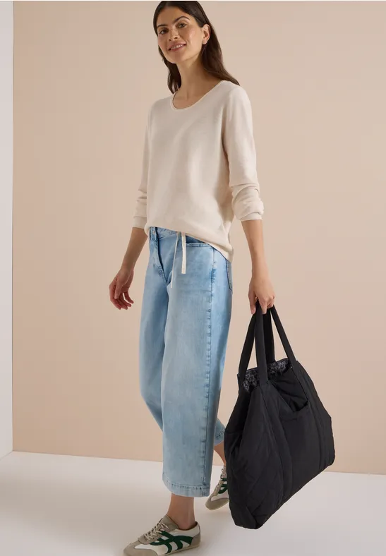 Frau in beigem Pullover, weiten Jeans und Sneakers trägt große, schwarze Stepp-Tasche.