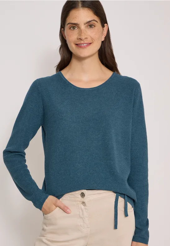 Frau mit blaugrünem Pullover und beigen Hosen, lächelnd vor hellem Hintergrund.
