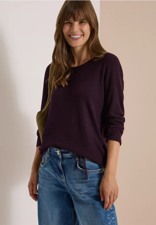 Frau mit lila Pullover und Jeans lächelt vor neutralem Hintergrund.