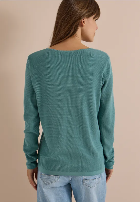 Une femme de dos porte un pull turquoise et un jean devant un fond uni.