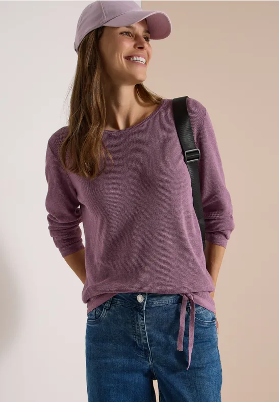 Jeune femme souriante en jean, haut violet, casquette et sac à bandoulière.
