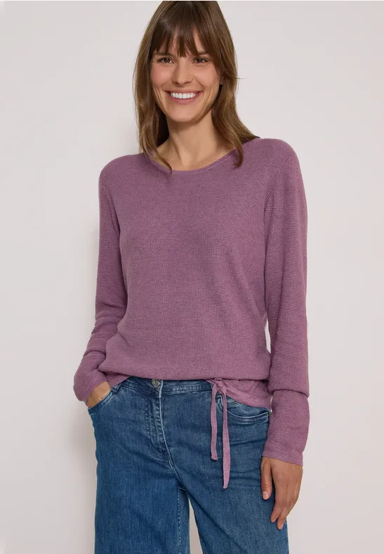 Une femme souriante porte un pull violet et un jean bleu devant un fond clair.