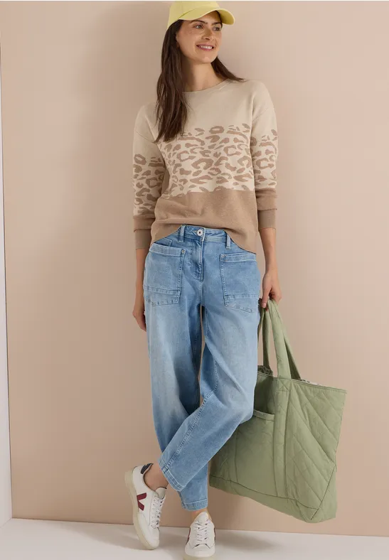 Model trägt beigefarbenen Pullover mit Muster, weite Jeans, weiße Sneaker und grüne Stofftasche.