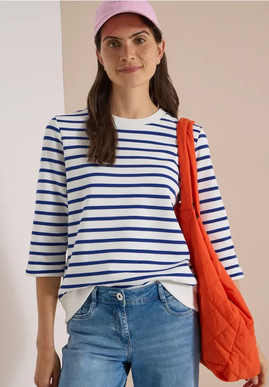 Femme portant un tee-shirt rayé bleu et blanc, un jean, une casquette rose et un sac orange.