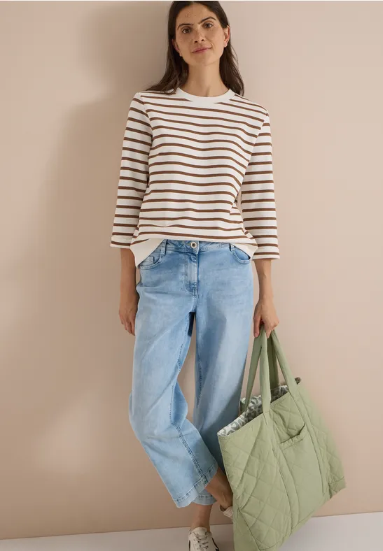 Frau mit weiß-blau gestreiftem Langarmshirt, hellblauen Jeans und großer grüner Stepp-Tasche vor neutralem Hintergrund.