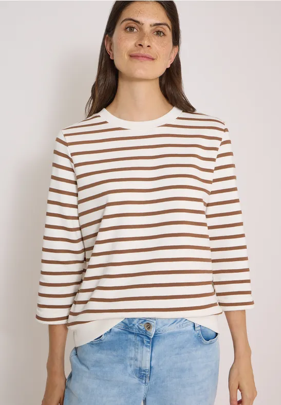 Frau in weiß-braun gestreiftem Shirt und blauen Jeans, frontal stehend, Haare offen, dezentes Lächeln.