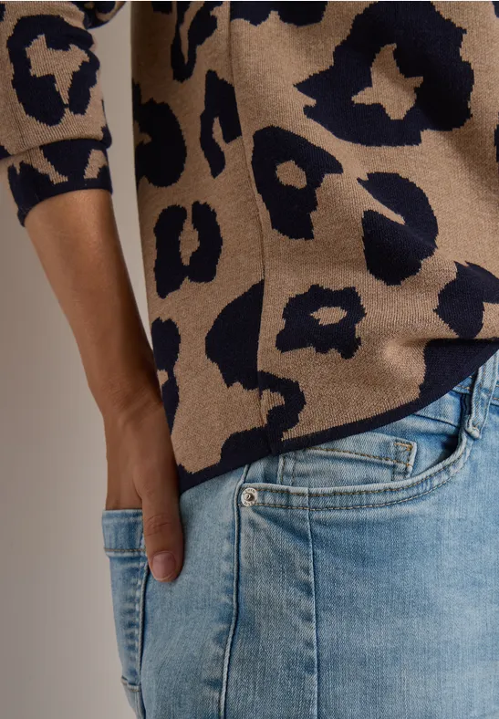Persoon in beige-blauwe trui met luipaardprint en lichtblauwe spijkerbroek, hand in zak