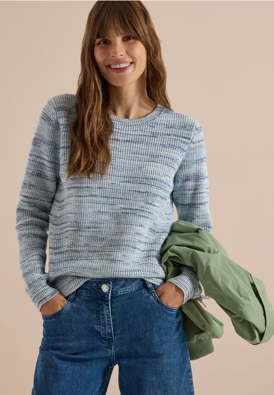 Femme souriante en pull bleu-gris, jean et veste verte devant un fond neutre.