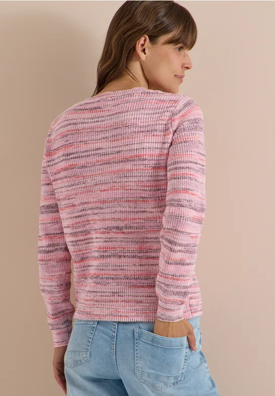 Une femme de dos porte un pull rose à rayures multicolores et un jean, les mains dans les poches.