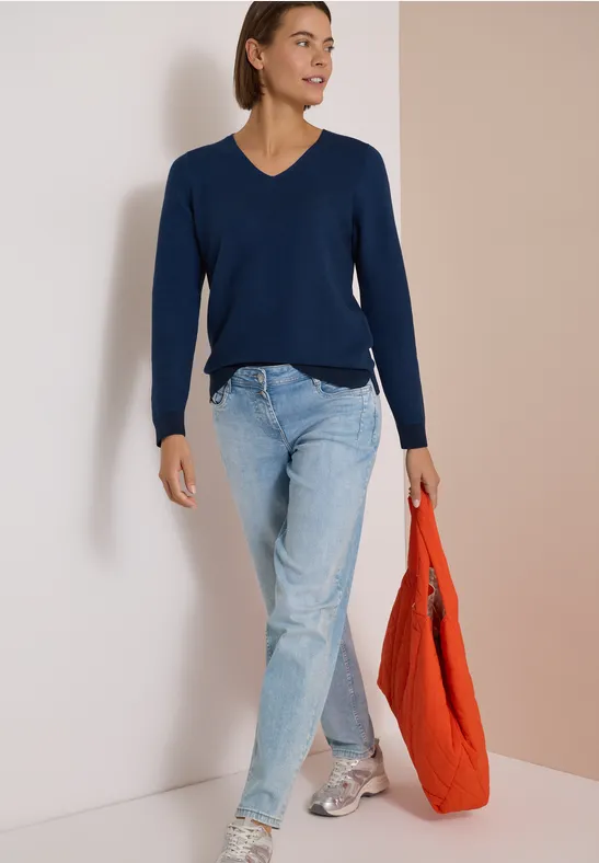 Femme en pull bleu marine à col V, jean clair et baskets, portant un sac orange.