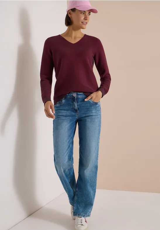 Femme casquette rose, pull framboise, jean large et baskets blanches appuyée contre un mur.