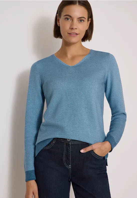 Femme en pull bleu à col V et jean, posant devant un fond blanc, une main dans la poche.