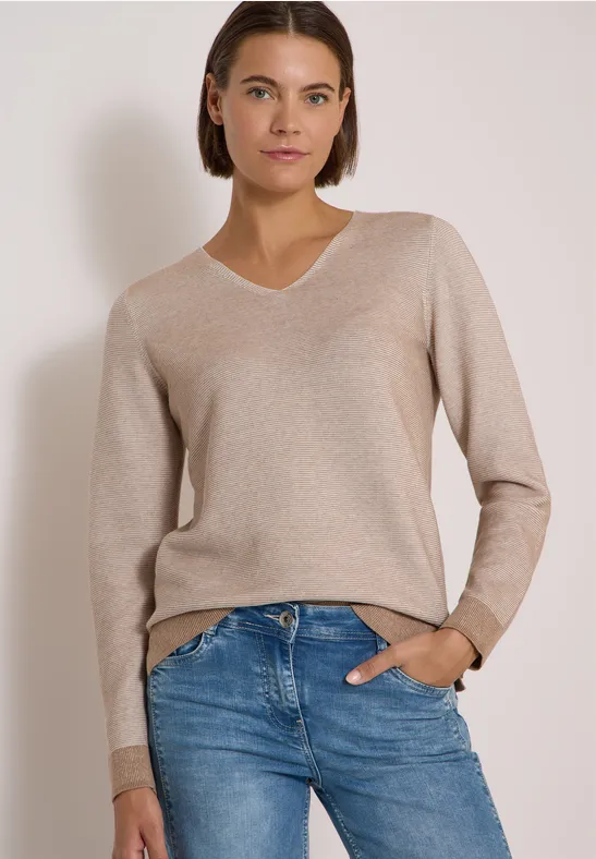 Une femme aux cheveux courts bruns porte un pull beige à col V et un jean bleu clair devant un fond uni.
