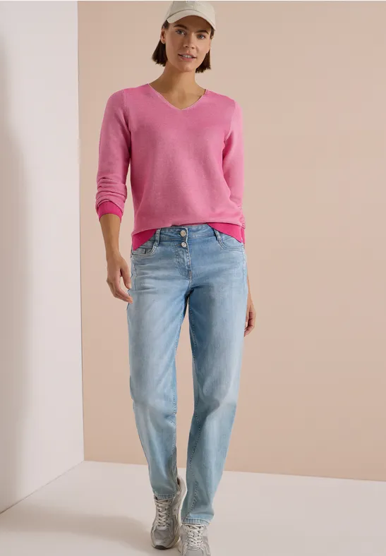 Femme en pull rose, jean et casquette blanche posant devant un mur beige.