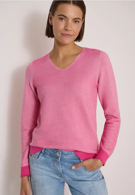 Femme en pull rose à col V et jean bleu, main dans la poche, fond clair.