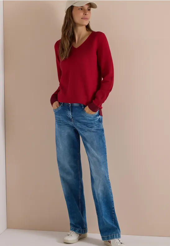 Femme en jean, pull rouge col V, casquette blanche et baskets devant un mur beige.