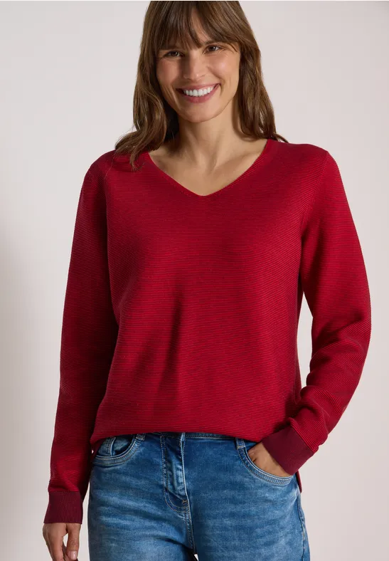 Femme souriante portant un pull col V rouge et un jean devant un fond clair.